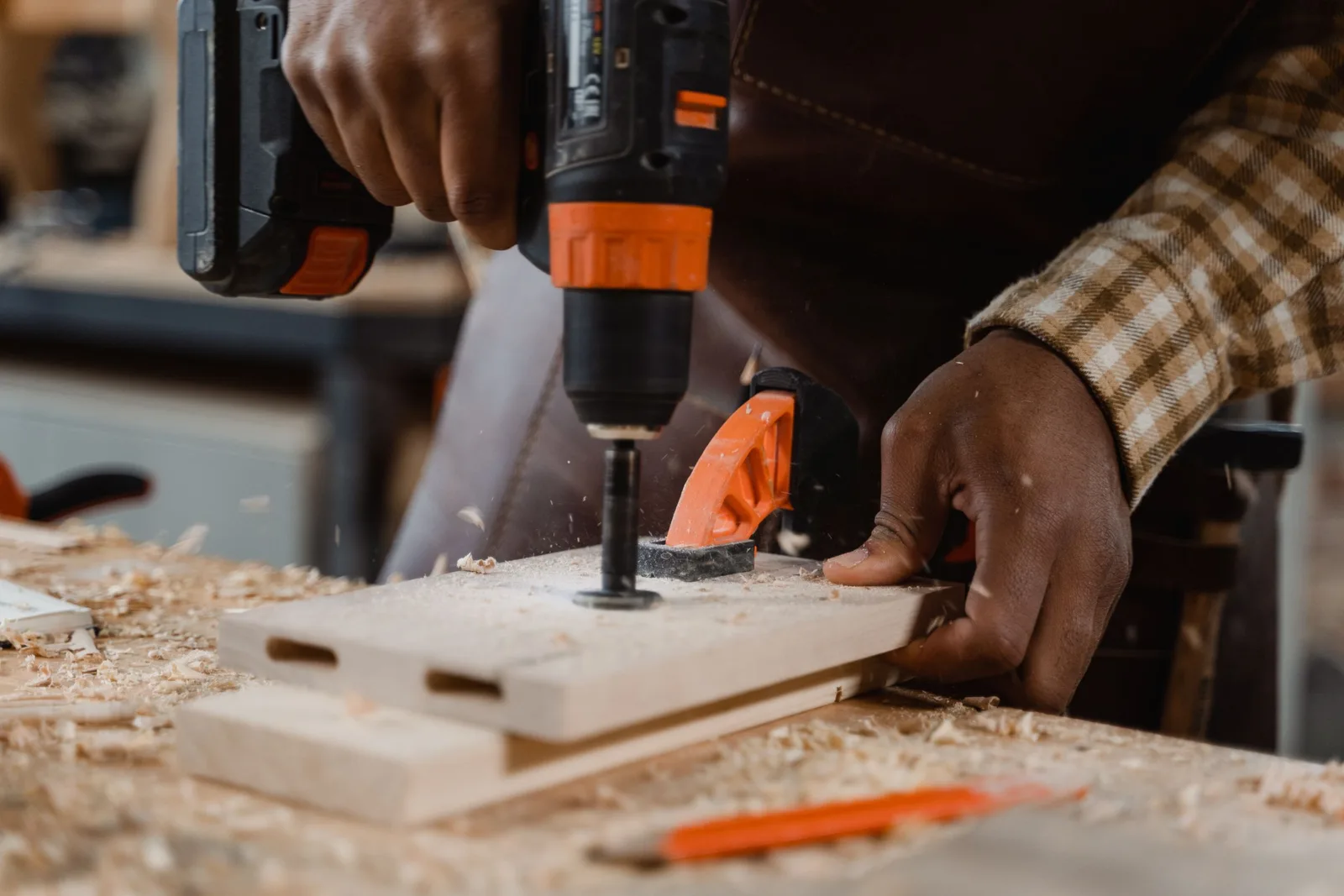 Une personne en tenue de bricolage utilise une perceuse sur un plan de travail en bois, dans un atelier clair.